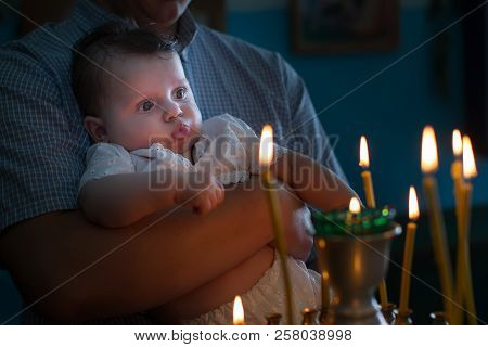The Father's Hands Hold The Baby In His Arms In The Church. The Kid Looks At The Candles Burning In 