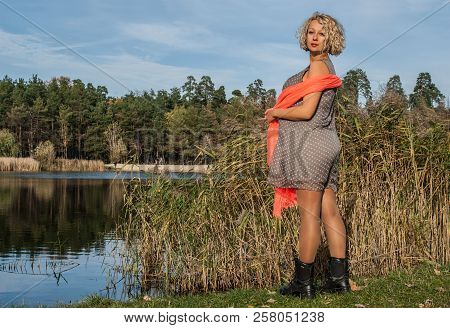 Pretty Pregnant Woman In Bright Orange Scarf Standing In Half Turn Near Blue Lake Of Autumn Park. Co