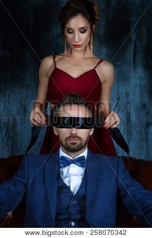 Couple In Dating. Woman Female In Expensive Red Evening Dress Tying Woman Eyes.  Rich Man Male Dress