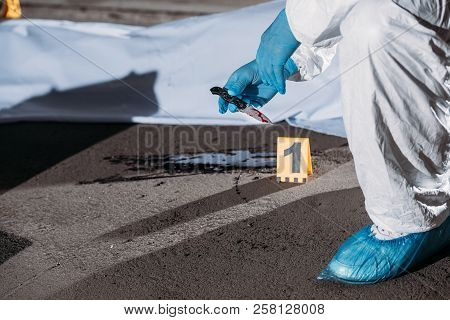 Partial View Of Criminologist In Latex Gloves And Protective Suit Holding Knife Above Blood On Groun