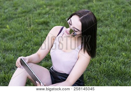 Full-length Portrait Of Young Businesswoman Sitting On Fresh Grass And Using Tablet Pc. Brunette Fem