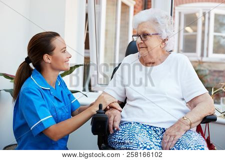 Senior Woman Sitting In Motorized Wheelchair Talking With Nurse In Retirement Home