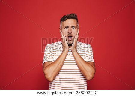 Portrait of uptight nervous man grabbing his face in confusion and stress isolated over red background