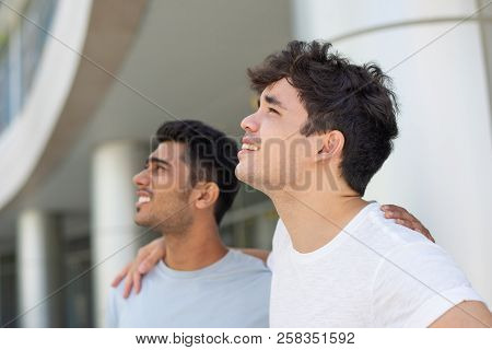Portrait Of Cheerful Male Friends Embracing And Looking Up Outdoors. Young Indian And Hispanic Male 
