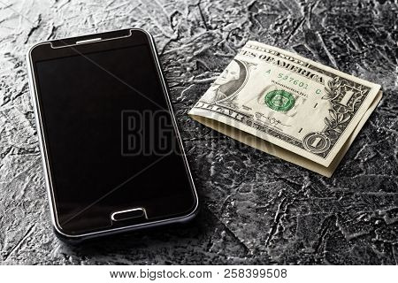 Phone And One Dollar On A Dark, Heterogeneous Background. Black, Brown, Gray, Color.