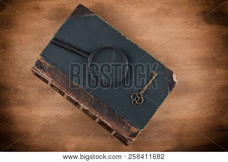 Old Book, A Key And A Magnifying Glass On The Book Against A Worn Wooden Background With Dark Edges 