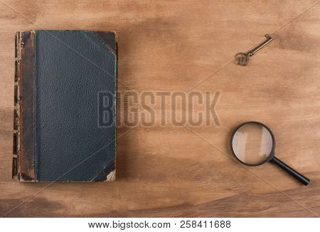 Old Book, A Key And A Magnifying Glass Against A Worn Wooden Background (top View, Copy Space In The