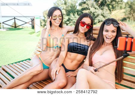 Very Nice Young Women Sits On Sunbed And Pose. Model On The Right Holds Phone On Stick. She Takes Se