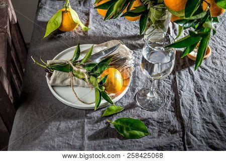 Table Setting With White Plate, Cutlery, Linen Napkin And Orange Tree Branch Decoration On Gray Line