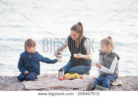 Happy Family In Warm Sleeveless Jackets Sitting On Sea Beach On Picnic With Tea Fruits