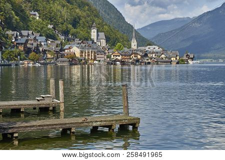 Hallstatt Village Landscape With Old Buildings Reflected In Blue Lake With Wooden Decks In The Foreg