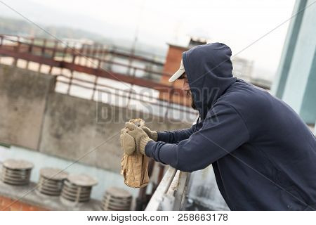 Homeless Drunkard Standing On A Building Rooftop Terrace, Wearing A Hoodie And Gloves, Holding A Bot