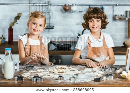 Cute Happy Kids In Aprons Preparing Tatsy Cookies And Smiling At Camera