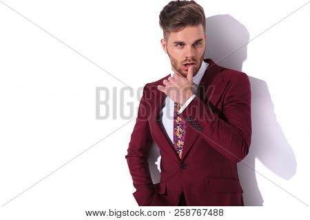 sensual portrait of a handsome elegant man in burgundy suit and tie, touching his lips and looking away to side on white wall