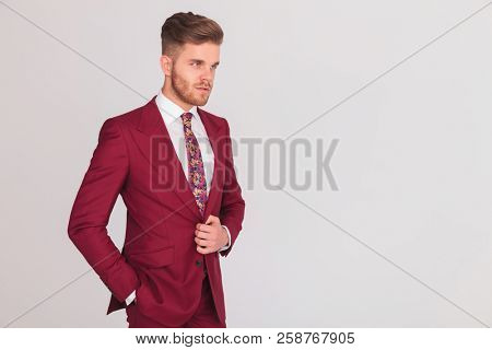 side view portrait of a relaxed modern business man standing on grey background with one hand in pocket and the other holding his suits's button