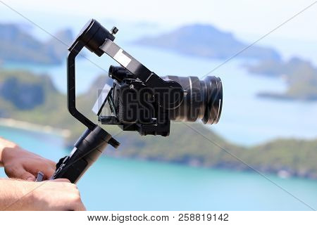 Photographer Holding  The Camera Gimbal Stabilizer On Top Of The Mountain. Ocean And Blue Sky Backgr