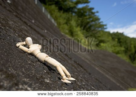 Wooden Doll Sleeping Face Up On A Concrete Slope In Front Of Green Forest Under Blue Sky