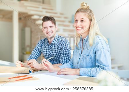 Happy young blond woman in denim casualwear and her colleague sitting by table in sudio and getting new ideas for project