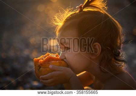 A Little Girl Is Eating A Peach And Is Illuminated By The Golden Light Of The Evening Sun. Girl Bite