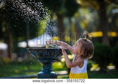 A Little Girl Of 3 Years Old Plays On The Street With A Stream Of Drinking Fountain And Enjoys The S