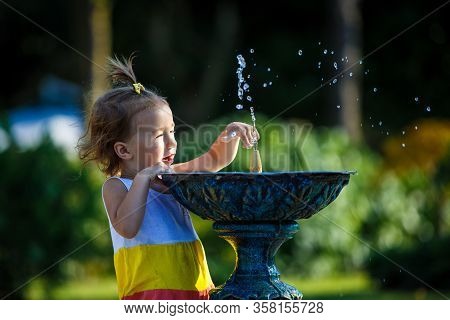 A Little Girl Of 3 Years Old Plays On The Street With A Stream Of Drinking Fountain And Enjoys The S