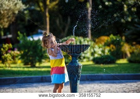 A little girl of 3 years old plays on the street with a stream of drinking fountain and enjoys the s