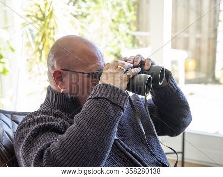 Elderly Man Sittiing, Looking Through A Glass Door With Binoculars As He Watches Something In The Di