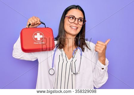 Young beautiful brunette doctor woman wearing stethoscope and coat holding first aid kit pointing thumb up to the side smiling happy with open mouth