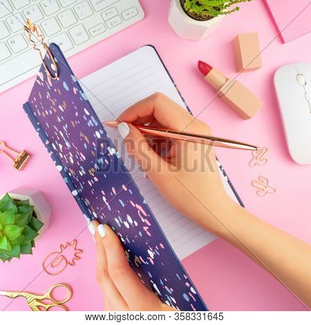 Womans Hands With White Manicure Writes In A Blue Notebook With A Pen. Work Place On Pink Background