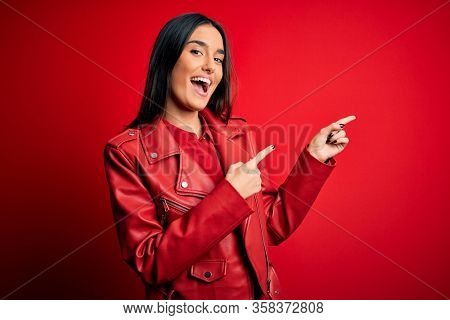 Young beautiful brunette woman wearing casual jacket standing over isolated red background smiling and looking at the camera pointing with two hands and fingers to the side.