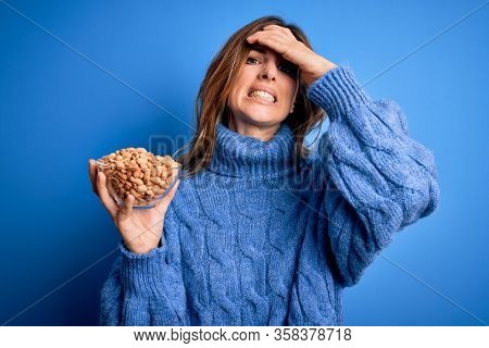 Young beautiful brunette woman holding bowl with peanuts over blue background stressed with hand on head, shocked with shame and surprise face, angry and frustrated. Fear and upset for mistake.