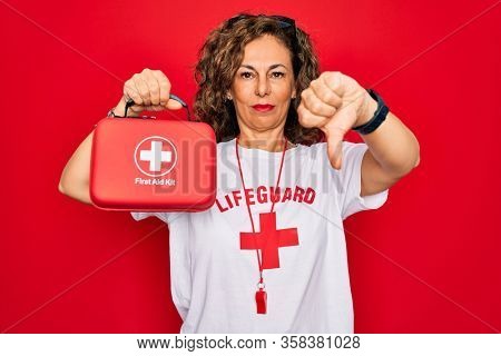 Middle age senior summer lifeguard woman holding first aid kit over red background with angry face, negative sign showing dislike with thumbs down, rejection concept