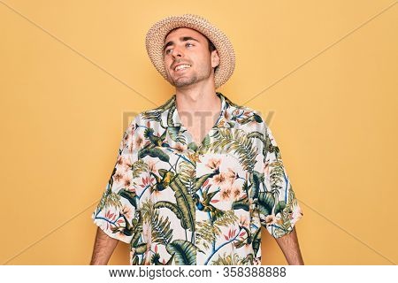 Young handsome man with blue eyes on vacation wearing summer florar shirt and hat looking away to side with smile on face, natural expression. Laughing confident.