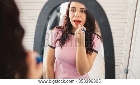 Young Beautiful Woman In Pajamas Paints Her Lips Of Red Lipstick, Looking In Mirror At Home. Close U