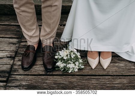 Legs Of Newlyweds On Wooden Background. Stylish Shoes Of Bride And Groom Outdoors. Bridal Bouquet On