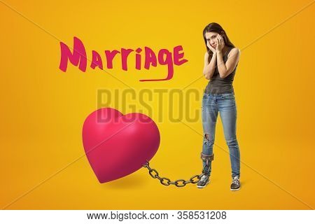 Young Frustrated Girl In Casual Clothes With Her Foot Chained To Big Cute Pink Heart On Amber Backgr