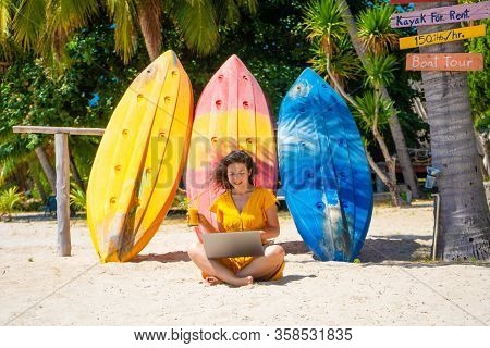 Girl In A Yellow Dress On A Tropical Sandy Beach Works On A Laptop Near Kayaks And Drinks Fresh Mang