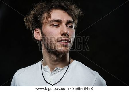 Curly And Hansome Caucasian Man Standing In A Dark Studio On A Grey Background, Wearing Casual White