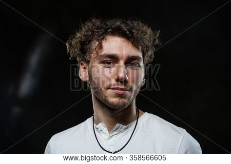Curly And Hansome Caucasian Man Standing In A Dark Studio On A Grey Background, Wearing Casual White
