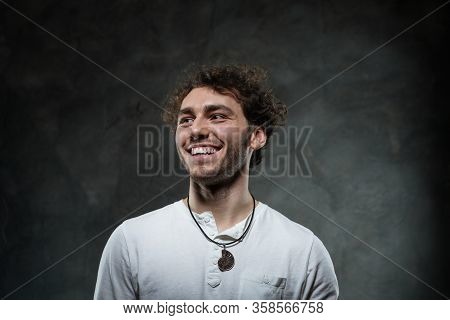 Curly And Hansome Caucasian Man Standing In A Dark Studio On A Grey Background, Wearing Casual White