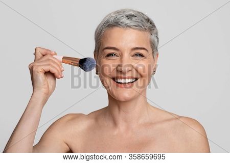 Portrait Of Beautiful Smiling Middle-aged Woman Doing Makeup, Applying Blush To Her Cheek And Lookin