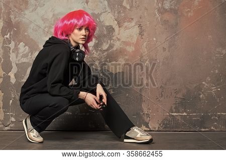 Girl In Sport Suit Sit On Hunkers On Abstract Wall. Woman Wear Pink Wig Hair With Headphones. Fashio