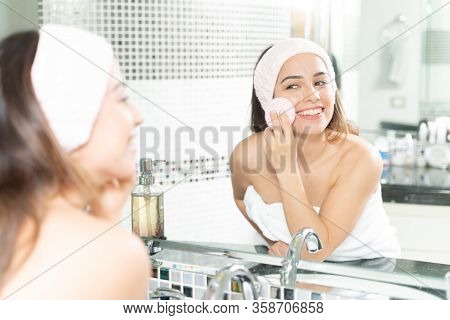 Pretty Woman With Headband Using A Sponge To Scrub Her Face While Getting Ready In The Bathroom