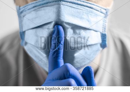 A Close-up Photograph Of A Hand In A Protective Medical Glove Shows A Hush Gesture Against A Face In