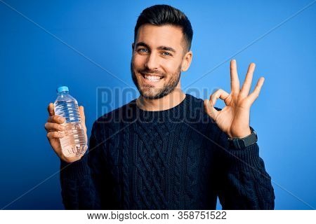 Young handsome man drinking bottle of water to refeshment over blue background doing ok sign with fingers, excellent symbol