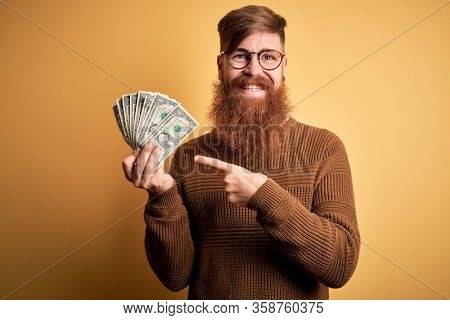 Redhead Irish man with beard holding a bunch of 1 dolla banknotes over yellow background very happy pointing with hand and finger