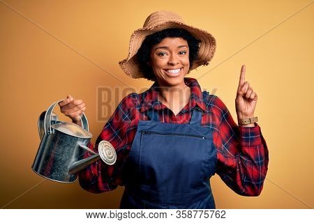 Young African American farmer woman with curly hair wearing apron and hat using water can surprised with an idea or question pointing finger with happy face, number one