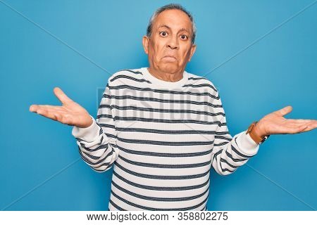 Senior handsome grey-haired man wearing striped sweater over isolated blue background clueless and confused expression with arms and hands raised. Doubt concept.