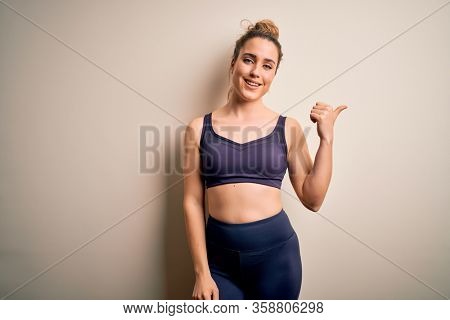 Young beautiful blonde sportswoman doing sport wearing sportswear over white background smiling with happy face looking and pointing to the side with thumb up.