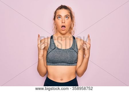 Young beautiful blonde sportswoman with blue eyes doing exercise wearing sportswear amazed and surprised looking up and pointing with fingers and raised arms.